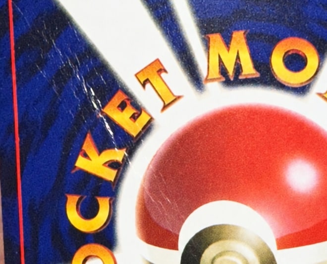 Close-up of the left side of a Pocket Monsters card back before the crease was fixed: a white crease line runs diagonally through the blue and across the orange lettering beside the Poké Ball.